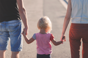 The backs of a mother and father holding the hands of their toddler child.