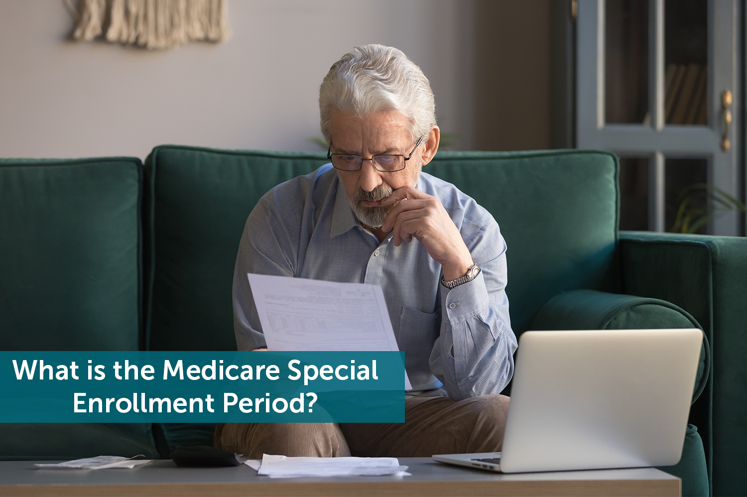 Elderly man sitting on a green couch looking at a piece of paper in front of an open laptop, wondering how Medicare Special Enrollment Period works.